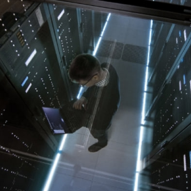 Overhead view of an engineer with a laptop walking between lit server racks.